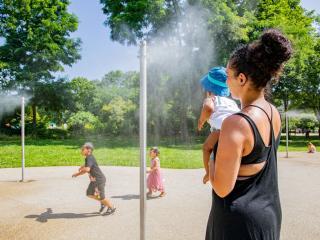 Photographie où l'on voit des brumisateurs vaporisent de l'eau dans le parc du Cerisier un enfant court vers eux, une autre marche, tandis qu'une mère de famille approche prudemment avec son bébé. - Agrandir l'image, fenêtre modale